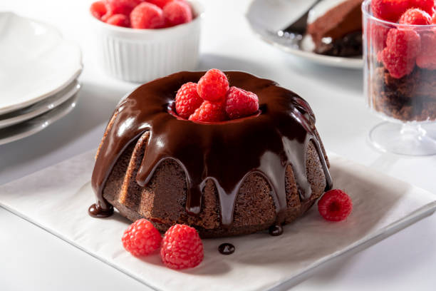 Chocolate Bundt Cake with Chocolate Ganache Beautiful mini chocolate bundt cake with chocolate ganache and raspberries. The delicious cake sits on a white table with plates, dish of raspberries and other items in the background out of focus. chocolate cake stock pictures, royalty-free photos & images