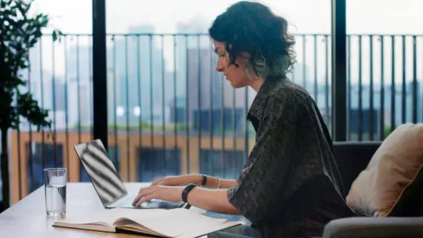 Stock image of a dark-haired young woman working in a high rise city apartment on her laptop.