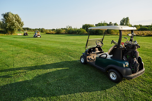 Golf car with golf clubs and equipment stands on grass golf course with long shadow during golfing
