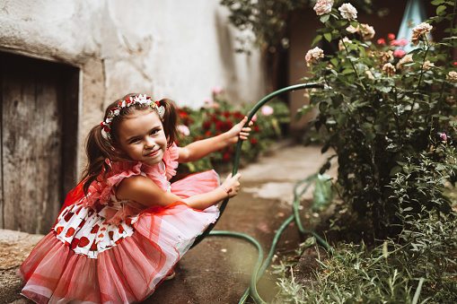 Cute Female Child In Pink Dress Watering Flowers With Hose In Backyard