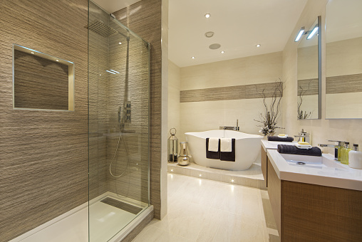 an elegantly designed and dressed bathroom in an contemporary new home with a walk-in shower area to the left, an unusually shaped bathtub in the centre and 'his and hers' basin pedestals to the right.