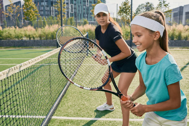 bambina positiva che tiene una racchetta mentre impara a giocare a tennis con la sua allenatrice sul campo all'aperto. istruttore individuale per un bambino per l'allenamento di tennis - tennis court tennis racket forehand foto e immagini stock