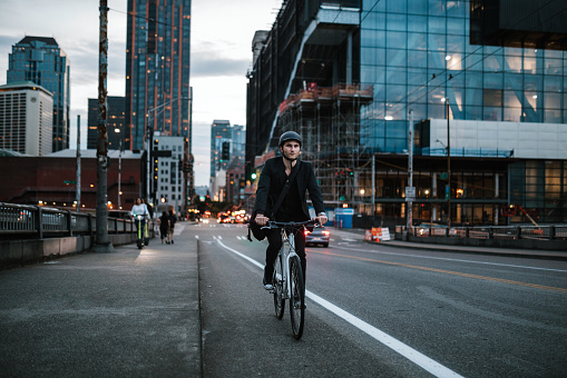 A Caucasian business man wearing a blazer rides his bicycle through the city streets of Seattle, Washington, USA.  He carries work items in a messenger bag slung over his shoulder.   An environmentally friendly and healthy way to commute to work: sustainable lifestyle.