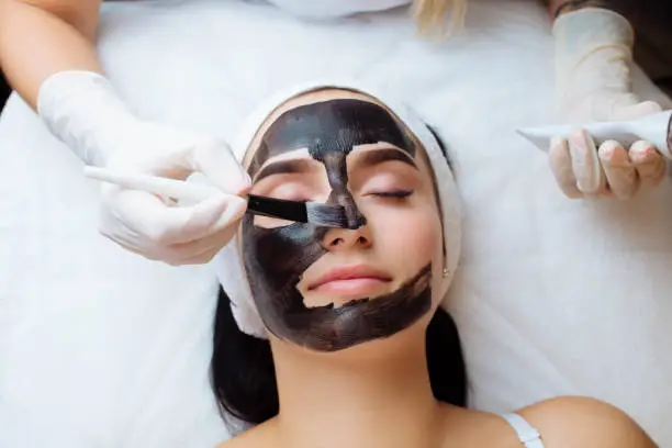 Photo of cosmetologist applying black mask on pretty woman face wearing black gloves, gorgeous woman in spa having facial procedures