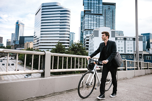 A Caucasian business man wearing a blazer walks his bicycle through the city streets of Seattle, Washington, USA.  He carries work items in a messenger bag slung over his shoulder.   An environmentally friendly and healthy way to commute to work: sustainable lifestyle.