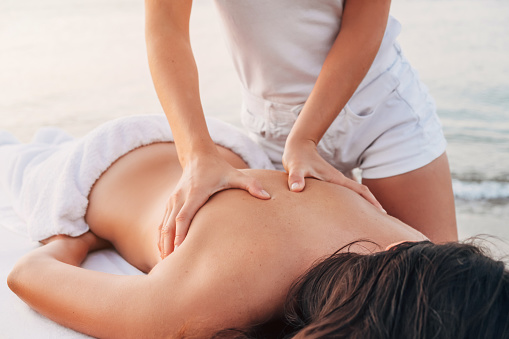 Girl masseur in white clothes on the seashore makes a back massage to a woman who lies on a massage table.