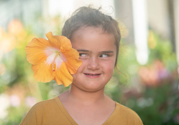 An adorable young girl around eight (8) years old has a silly expression on her face as she looks at the giant yellow hibiscus flower that was placed on her ear.