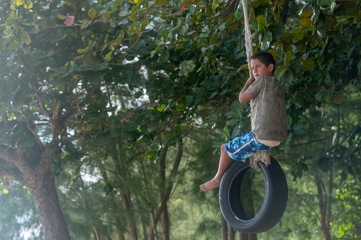 A young boy is hanging on a tire swing outdoors. There are big mature trees in the background. He has both hands holding onto the rope of the tire swing.