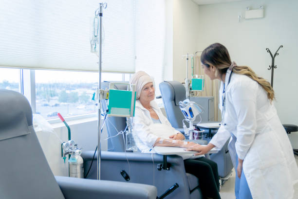 Senior woman with cancer talking to a female oncologist A senior woman is sitting on a chair in a chemotherapy treatment room. She is wearing a headscarf due to hair loss. Her female oncologist is checking in on her and providing her with moral encouragement. chemotherapy drug stock pictures, royalty-free photos & images