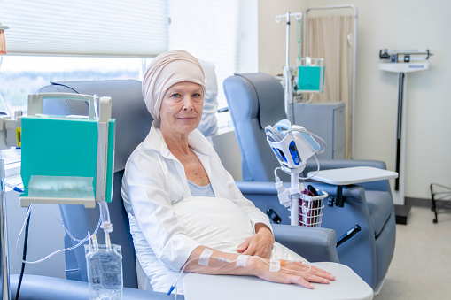 A portrait of a senior woman sitting on a chair in a chemotherapy treatment room. She is wearing a headscarf due to hair loss. She has a focused expression on her face and is looking at the camera.