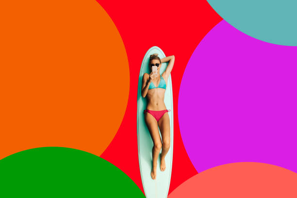 Surfing the internet and surfing the ocean Photo of a young woman enjoying while lying down on a surfboard and using smart phone; isolated on an colorful background. extreme sports technology stock pictures, royalty-free photos & images