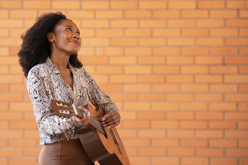 Cheerful afro american woman playing guitar outside looking up on a brick wall background. Selective focus.
