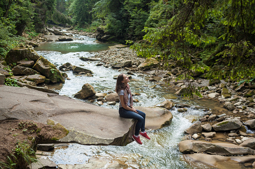 Happy smiling girl in a white t-shirt, blue jeans and red sneakers sitting near mountain river. Young woman enjoys fresh air in forest resting on a rock above a waterfall. lonely travel, side view