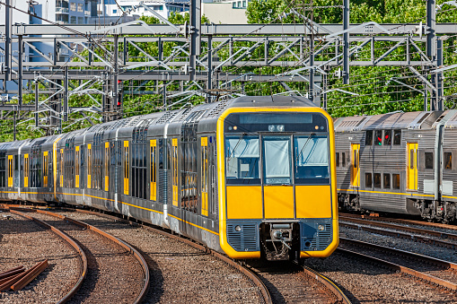 Modern double-deck (bi-level) electric commuter train with yellow front and doors passing an older  train also with yellow doors as they approach major city station: track is tree-lined. ID and logos edited. Yellow is dominant colour