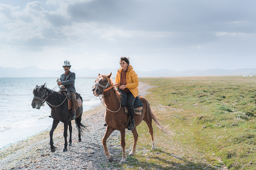 Young Caucasian woman in yellow down jacket  riding on horse near Son-Kul lake in Kyrygzstan with local guide