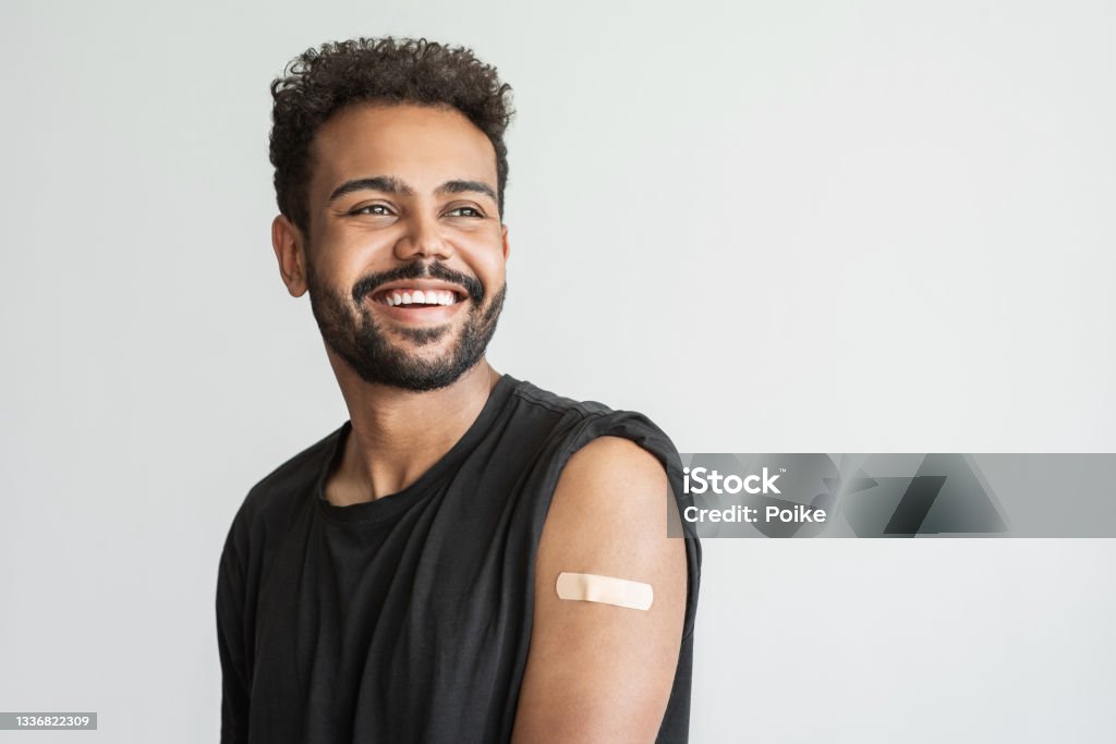 Man showing his vaccinated arm with plaster, got COVID-19 vaccine Young man received a corona vaccine looking away, isolated on gray background. Corona virus protection, self care, healthy lifestyle, vaccination concept. Vaccination Stock Photo