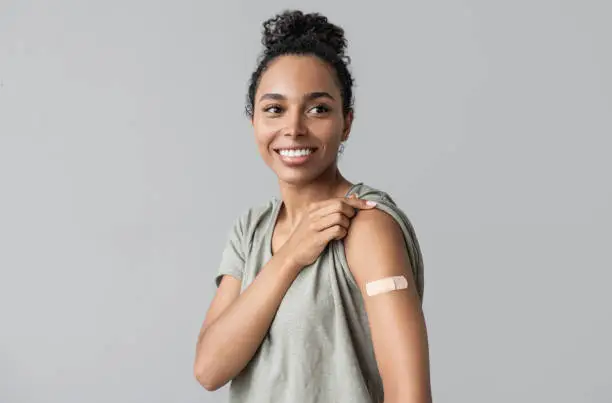 Smiling happy woman showing her arm with bandage after receiving covid-19 vaccination. Isolated on gray background. Corona virus protection, self care, healthy lifestyle concept.