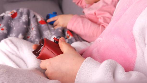 istock Happy Caucasian Kids Wearing Cosy Pyjamas Playing Cooperative Video Game using Handheld Gaming Console Controllers While Sitting On A Couch in a Living Room before Going to Bed Close Up 1336836249