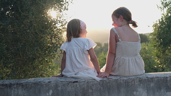 Two Young Caucasian Sisters Spending Time Togheter Talking Hugging and Laughing when Sitting on Tuscan Villa Terrace Wall During Golden Sunshine Afternoon