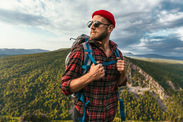 A bearded traveler with a backpack on the top of a mountain. Portrait of a traveler in a red cap and sunglasses. A tourist with a backpack stands against the background of a mountain. Copy space stock photo
