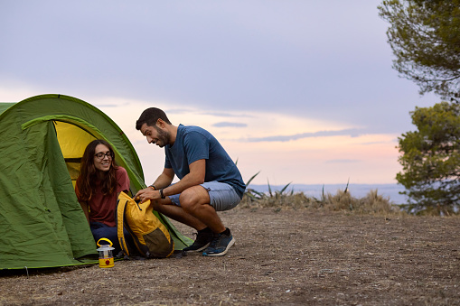 Smiling woman looking while man opening bag by tent on cliff. Male and female camping during vacation. They are spending leisure time together.