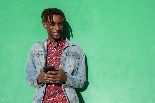 Young african man with smartphone laughing on green colored background.