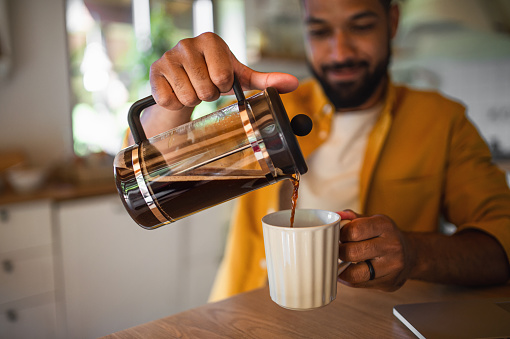 A young man pouring coffee from french press working indoors, home office concept.