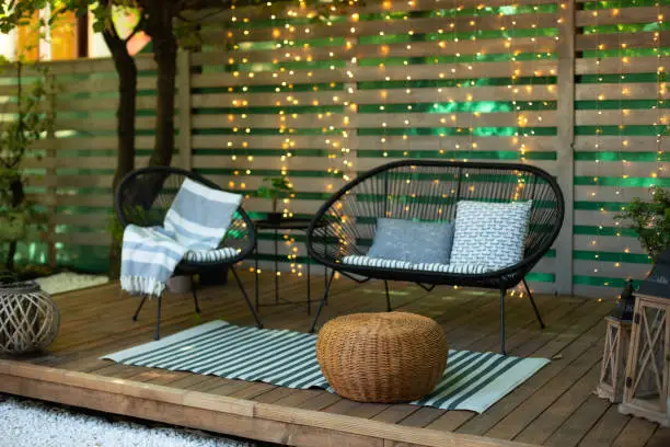 Photo of Terrace house with plants, wooden wall and table, comfortable sofa, armchair and lanterns. Cozy space in patio or balcony. Wooden veranda with garden furniture. Modern lounge outdoors in backyard
