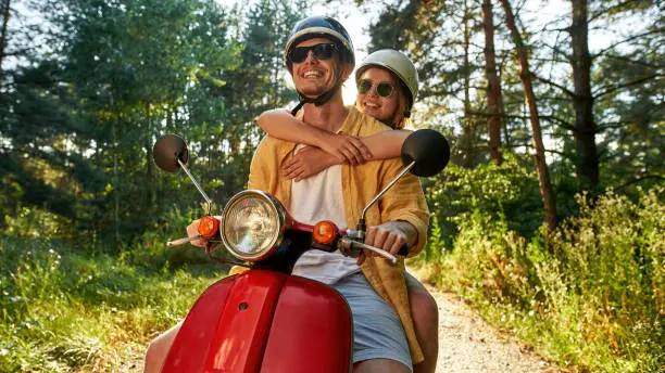 Smiling young caucasian friends wearing helmets and sunglasses riding scooter along road in sunny wood, widescreen. Leisure, fun and entertainment concept