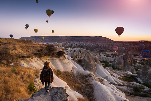 A young woman tourist in a hat standing on a mountain and looks at the sunrise and balloons in Cappadocia. Turkey. Top attraction travel destinations - Goreme National Park.