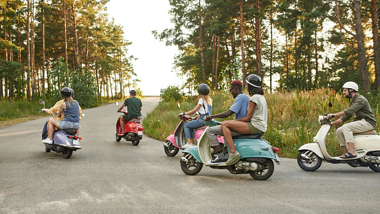 Company of young multiracial friends riding retro scooters along asphalt road in sunny nature, widescreen. Leisure, fun and entertainment concept