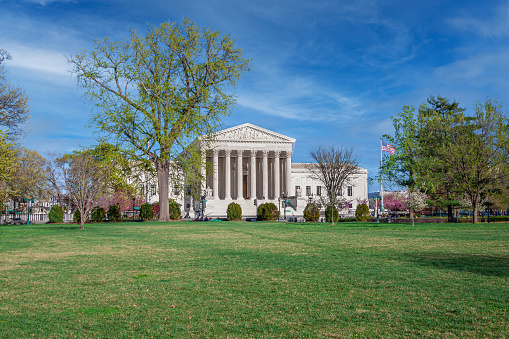 U.S. Supreme Court Building, Washington DC, USA. Green lawn, trees in bloom and bushes are in foreground. Blue sky with Puffy Clouds is in background. \n.