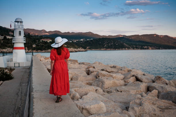 Beautiful picture of a  girl in a hat and red dress near the lighthouse and boats  at sunset. Port of Kas old town, Antalya, Turkey. stock photo