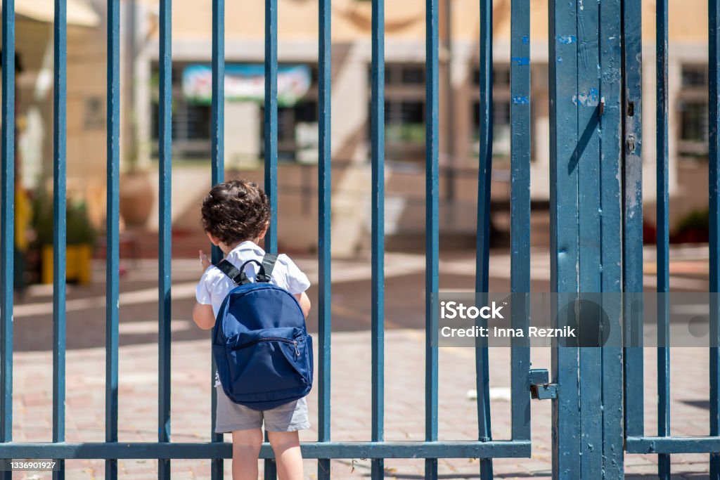 Child with bag back to school. Faceless cute little boy back to school with blue schoolbag Kid with school backpack look on schoolyard towards an open entrance or exit door. Schools and preschools remain locked for children during lockdown, coronavirus pandemic and second wave of covid-19. School Building Stock Photo
