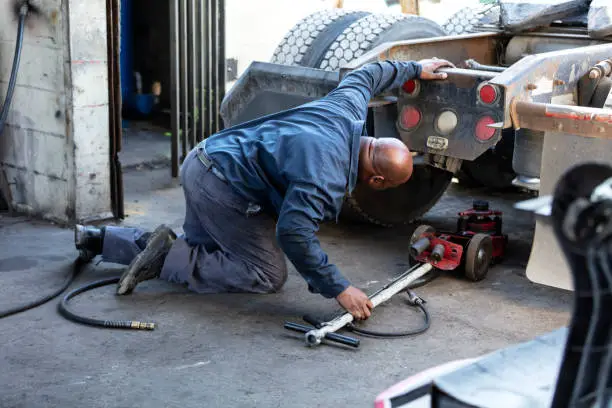 A mature African-American man in his 40s working in a truck repair shop. He is kneeling on the floor, looking under the rear of the truck, sliding a jack underneath.