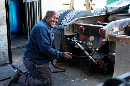 A mature African-American man in his 40s working in a truck repair shop. He is kneeling at the rear of truck with a jack, looking at the camera, smiling.
