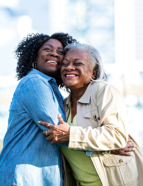 Two senior African-American women in city park, hugging Two senior African-American women in their 60s taking a walking together in a city park on the waterfront. They are best friends, hugging and laughing. cheek to cheek photos stock pictures, royalty-free photos & images