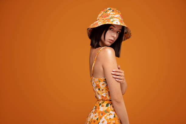 Fashionable young girl with natural freckles and colorful makeup wearing bucket hat , looking at camera, Fashionable young girl with natural freckles and colorful makeup wearing bucket hat , looking at camera, posing on orange studio background. A lot of copy space. Modern influencer woman. bucket hat stock pictures, royalty-free photos & images