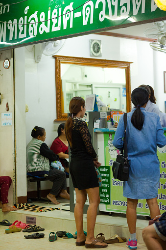 Thai people and women at pharmacy and doctor's office in Pak Chong in evening. Two young women are standing outside at reception. Many shoes are placed at entrance. Inside people are sitting and waiting. Pak Chong is in Nakhon Ratchasima province