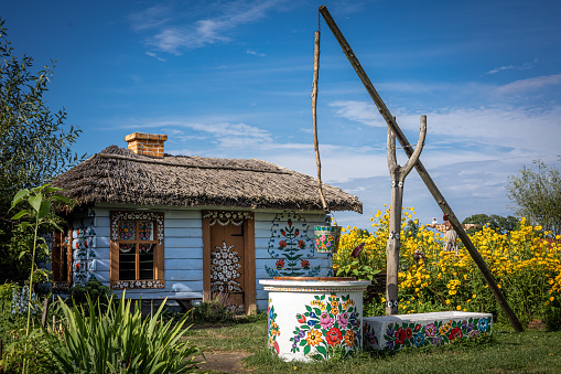 Zalipie, Poland - August 1, 2021: A small, white wooden cottage and a traditional well, painted in colorful flowery pattern. Lots of yellow flowers in the garden. Sunny, summer day.