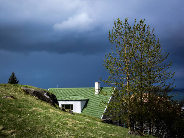 HAFNARFJORDUR, ICELAND - May 31, 2021: \nA scandinavian style house with green roof and chimney, hidden behind green hill. Dark, stormy sky. Hafnarfjordur, Iceland.