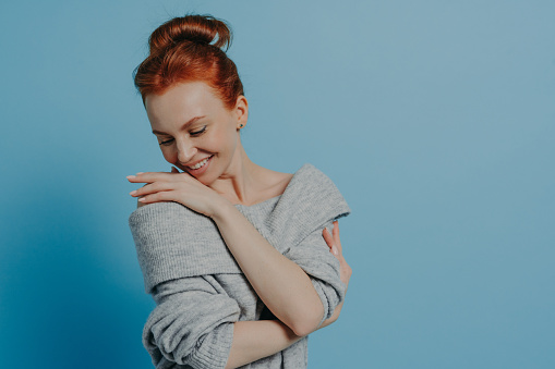 Young beautiful romantic red-haired woman wearing knitted sweater embracing herself with pleasure and smiling, feeling comfort while standing isolated over blue studio background