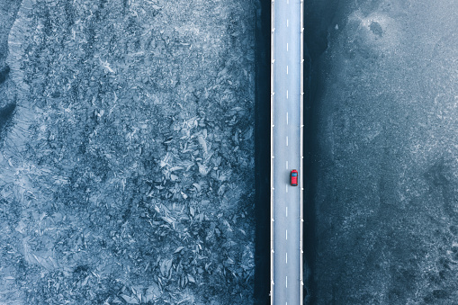 Aerial (drone) view on a red car driving over the bridge on a frozen lake.