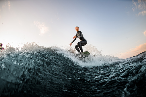 athletic female wakesurfer in wetsuit on the board rides down the river wave against the background of blue sky