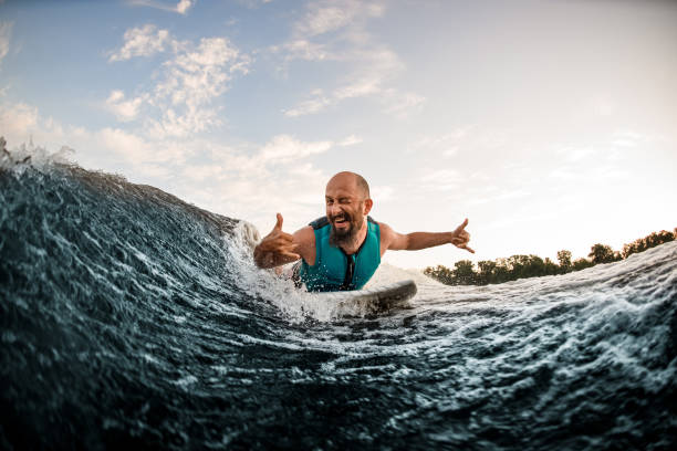 allegro wakesurfer maschio sdraiato a bordo cavalca giù onda e mostrare gesti e ammiccamenti delle mani - wakeboarding surfing men vacations foto e immagini stock