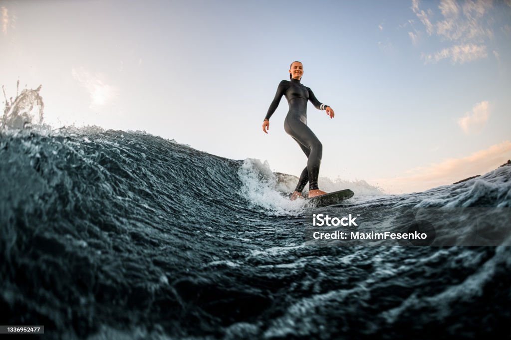 Young handsome woman wakesurfing on the board down the river against the background of blue sky Young handsome woman in wetsuit wakesurfing on the board and riding down the river wave against the background of blue sky Surfing Stock Photo