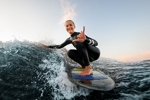 smiling woman in wetsuit sits on wakesurf board and rides the wave and touches the waves with one hand