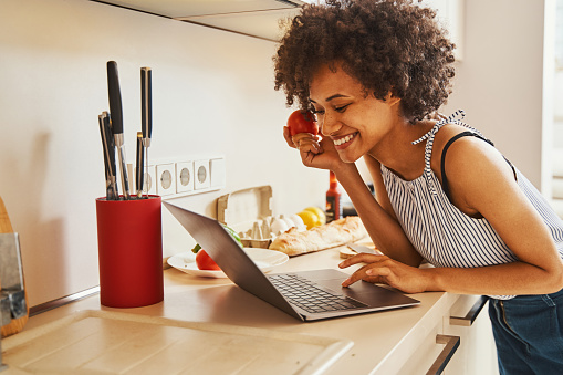 Smiling contented woman with a red tomato in one hand looking at the laptop screen