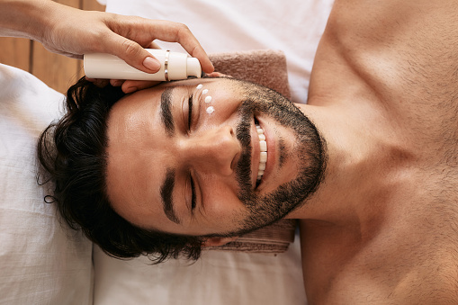 Beautician applies white skin moisturizer to handsome man's face at wellness center. Male patient enjoys beauty treatments, skincare and relaxation at spa