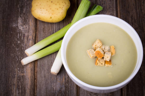A bowl of leek and potato soup with bread croutons, over old wood table with fresh leeks and potatoes alongside